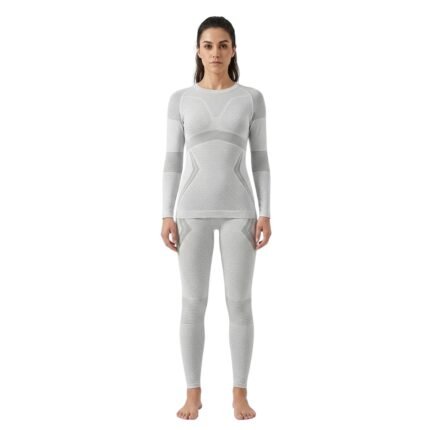 Smiling woman wearing a light grey seamless ski base layer set with long-sleeve top and leggings, standing front facing against a white background.