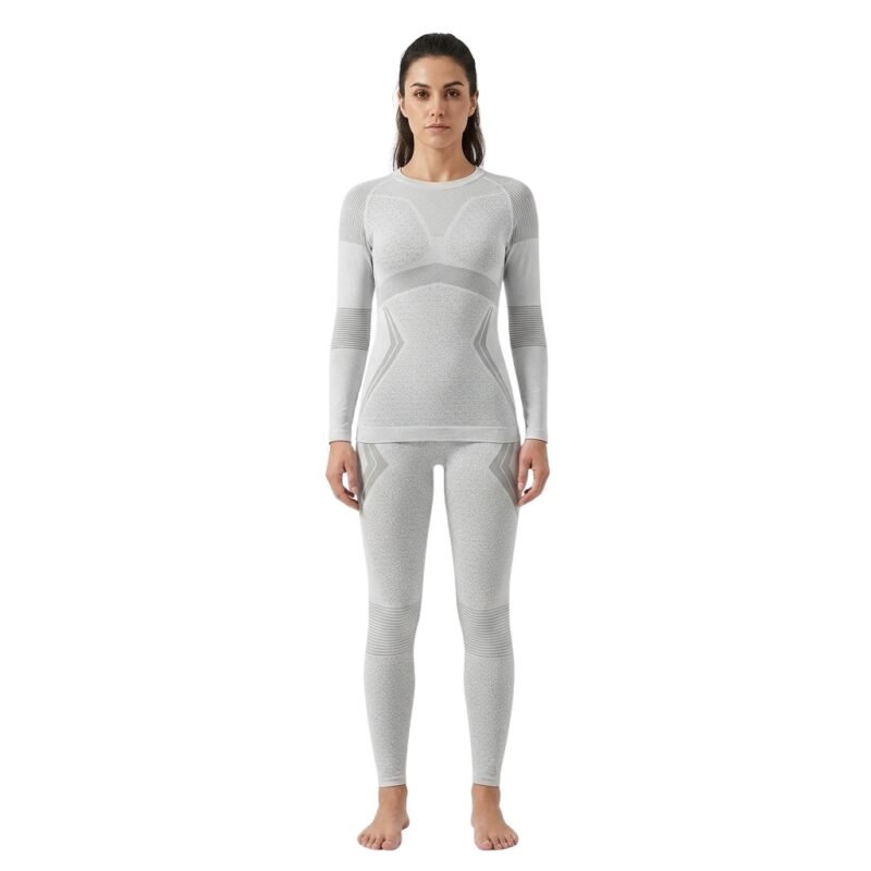 Smiling woman wearing a light grey seamless ski base layer set with long-sleeve top and leggings, standing front facing against a white background.