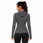 Back view of a female model wearing a custom gray compression hoodie by Eco Jersey, showing the attached hood, back panel construction, thumb holes, form-fitting silhouette, and black leggings