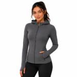Female model wearing a custom gray compression hoodie by Eco Jersey from the front, showing the full-zip closure, attached hood, thumb holes, side pocket, and body-contouring athletic fit with black leggings
