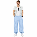 Smiling male model wearing custom sky blue pumper pants by Eco Jersey, styled with a white t-shirt, denim vest, and white sneakers, showcasing the wide balloon fit and relaxed jogger style