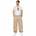 Smiling male model wearing custom tan beige pumper pants by Eco Jersey, styled with a white t-shirt, denim vest, and white sneakers, showcasing the relaxed balloon fit and casual streetwear style