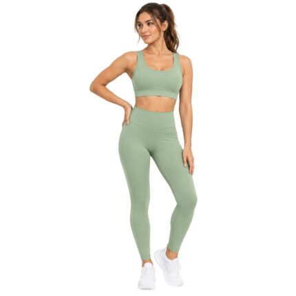 Front view of a fit woman in a light sage green sports bra and high-waisted leggings, hands on her hips, with her hair in a ponytail as she looks to the side.