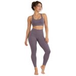 A woman with her dark hair in a bun wears a dusty lavender sports bra and matching high-waisted yoga leggings, standing on a plain white background.